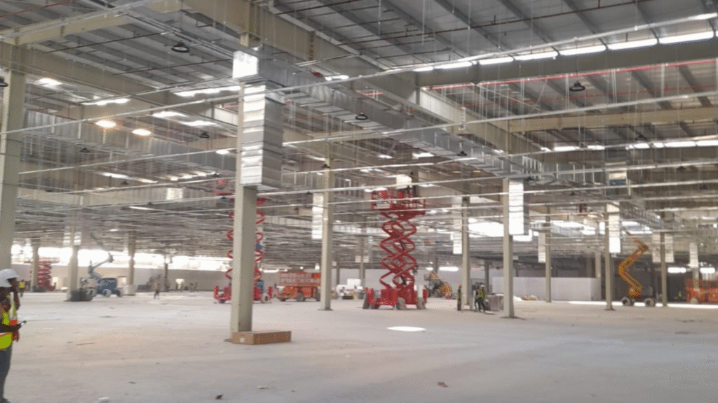 Interior of a large warehouse with workers performing plumbing installations using scissor lifts and other construction equipment.