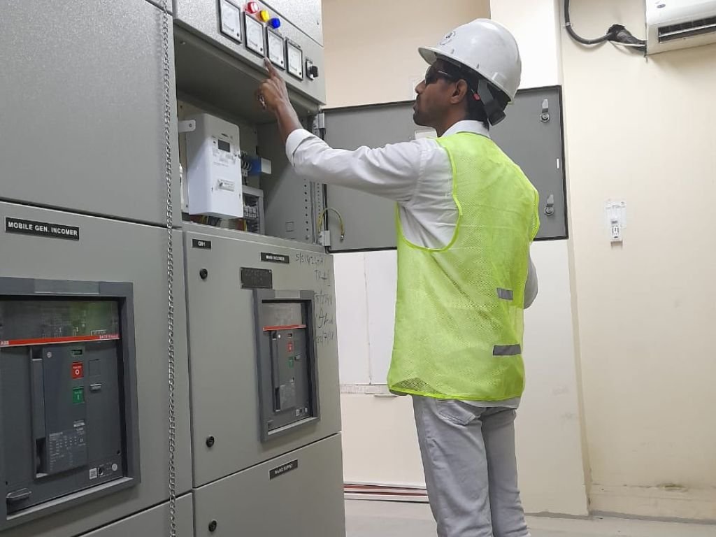 "Electrical engineer pptswearing safety helmet and reflective vest inspecting and adjusting electrical control panel in a utility room, performing electrical maintenance and testing in Sharjah, UAE."